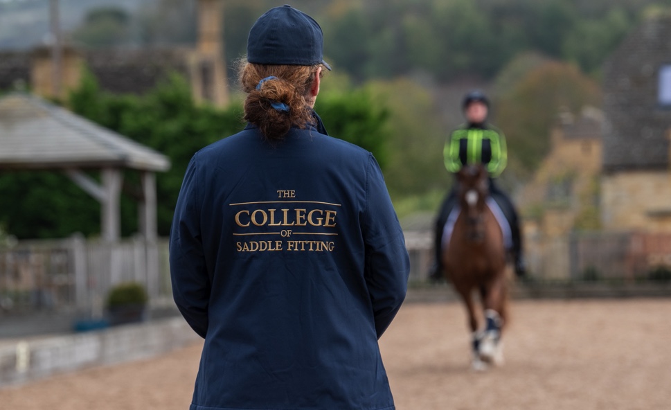 A new Saddle Fitting College Course will launch in February 2023 designed for equestrian professionals Image of lady watching a horse and rider with a college of saddle fitting branded jacket on.