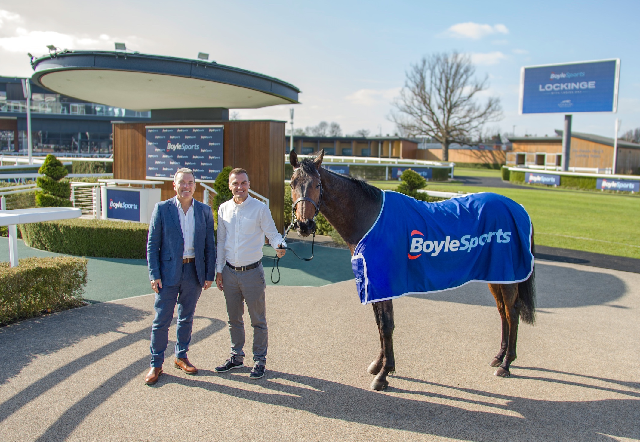 Horse stood in Boylesports rug at a racing track with two men